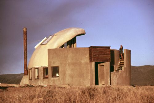 A unique, modern house with a curved roof and a person standing on stairs, set against a mountainous landscape.