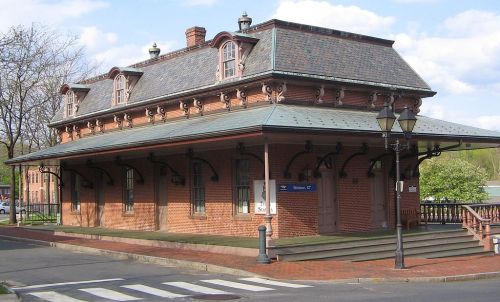 Historic brick train station with a sloped roof, decorative windows, and lampposts, set against a blue sky.