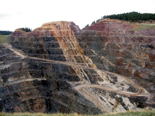 A large, multi-layered open-pit mine with steep, colorful rock formations and a winding road.