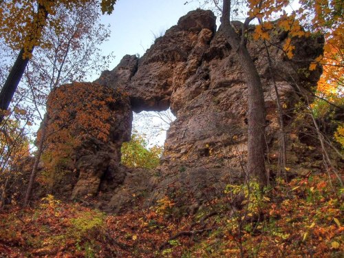 A rocky arch formation surrounded by trees and autumn foliage under a clear sky.