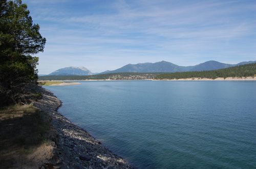 A serene lake surrounded by mountains under a clear blue sky, with a rocky shoreline and lush greenery.