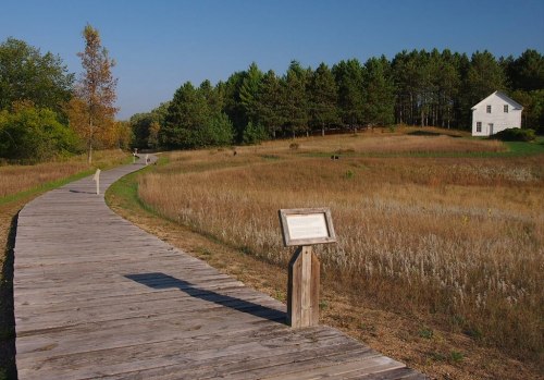 A wooden path leads through a grassy field to a white house, surrounded by trees under a clear blue sky.