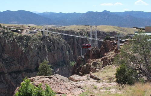 A cable car crosses a deep canyon with mountains in the background, surrounded by rocky terrain and greenery.