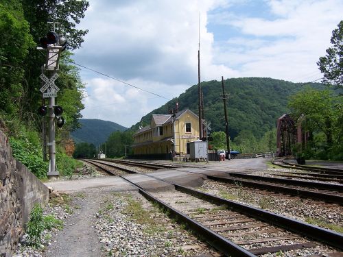 A yellow train station beside railroad tracks, surrounded by green hills and a cloudy sky.