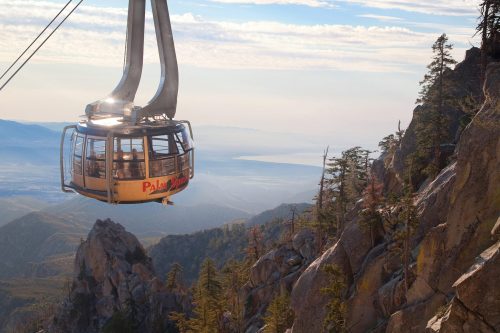 A gondola ascends a mountain, offering views of the valley and distant lake under a clear sky.