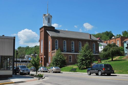 A brick church with a steeple stands beside a street lined with trees and parked cars under a blue sky.