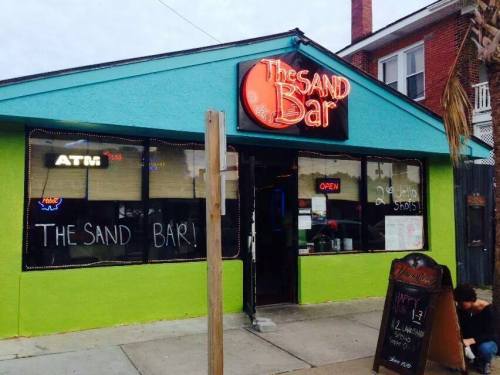 Exterior of The Sand Bar, a colorful building with a neon sign, open for business, featuring a chalkboard menu outside.