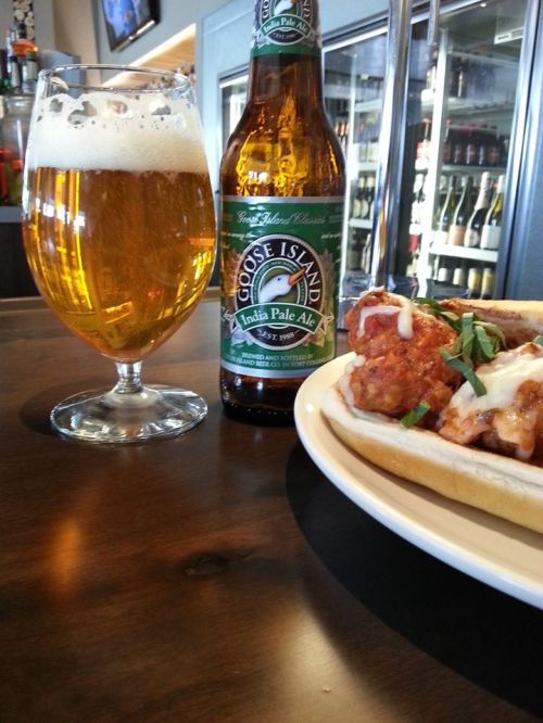 A glass of beer and a bottle of Goose Island IPA next to a plate of meatball sub topped with herbs.