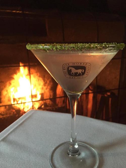 A martini glass with a green sugar rim sits on a table, with a warm fireplace glowing in the background.
