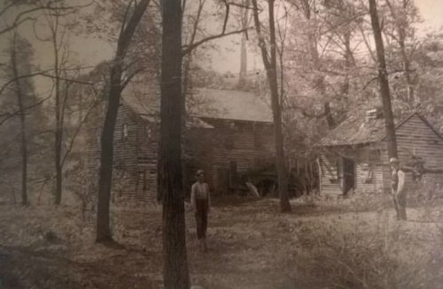 Black and white photo of two men standing near rustic wooden houses surrounded by trees.