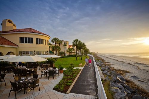A scenic beachfront view with a restaurant patio, palm trees, and a couple walking along the shore at sunset.