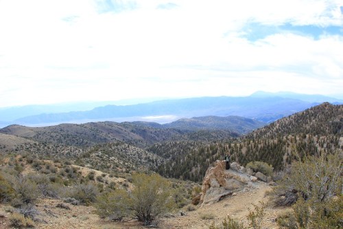 A person stands on a rocky outcrop overlooking a vast mountainous landscape under a cloudy sky.