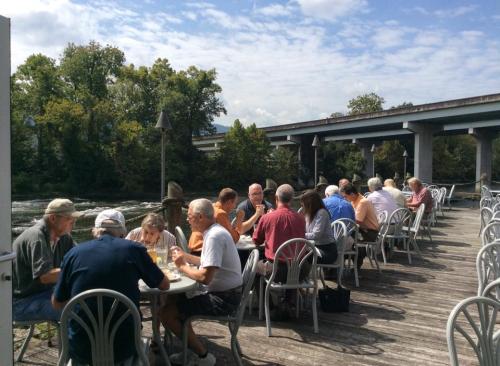 A group of people dining outdoors by a river, seated at tables along a wooden deck under a cloudy sky.