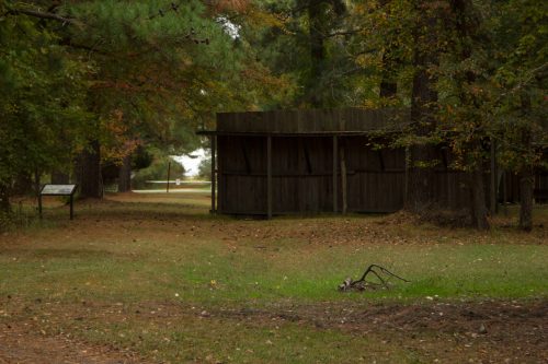 A rustic wooden structure surrounded by trees and a grassy area, with a path leading into the distance.