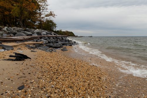 A rocky beach with pebbles and sand, gentle waves lapping at the shore under a cloudy sky.