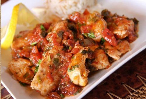 A plate of chicken in a tomato sauce, garnished with herbs and a lemon wedge, served with rice.