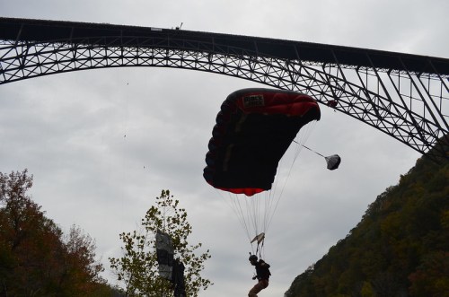A parachutist descends beneath a large bridge, surrounded by trees and a cloudy sky.