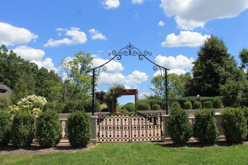 A decorative wrought iron gate opens to a lush garden under a bright blue sky with fluffy clouds.