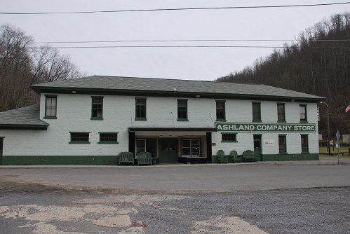 A green and white building labeled "Ashland Company Store" with a gravel parking area and mountains in the background.