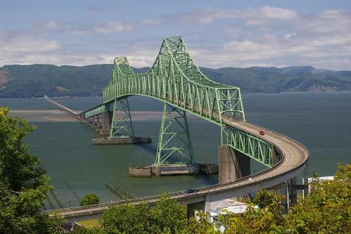 A green steel bridge spans a river, connecting land on either side with mountains in the background under a blue sky.