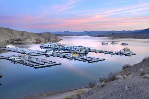 A serene marina at sunset, with boats docked and mountains in the background reflecting on calm waters.