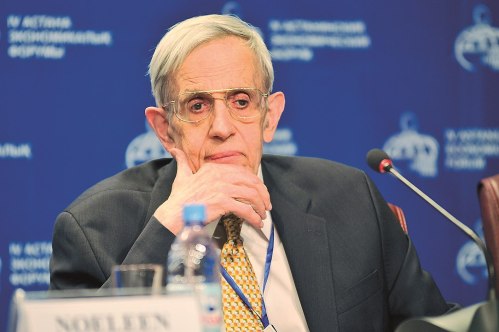 An elderly man with glasses sits thoughtfully at a conference, with a blue backdrop and a water bottle in front of him.