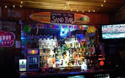 A vibrant bar scene featuring a colorful display of liquor bottles, neon lights, and a welcoming sign for the Sand Bar.