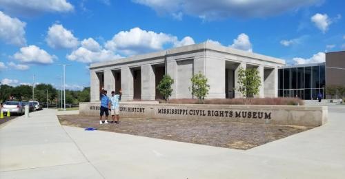 Two people stand in front of the Mississippi Civil Rights Museum, with a clear blue sky and clouds above.
