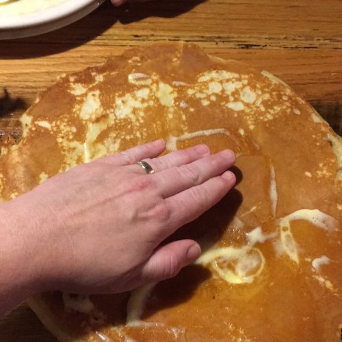 A hand rests on a large, golden-brown pancake on a wooden table.