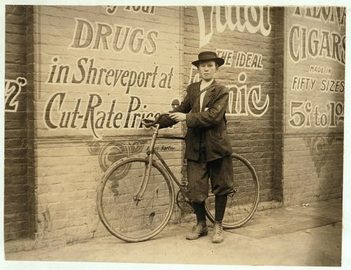 A young boy stands with a vintage bicycle in front of a wall covered in advertisements.