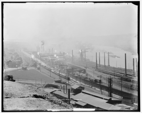 A black and white aerial view of an industrial area with smokestacks, trains, and a river in the background.