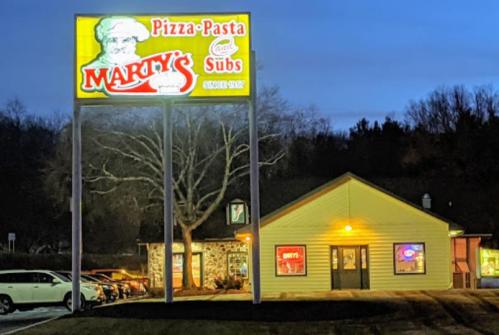 A restaurant sign for Marty's Pizza-Pasta-Subs illuminated at night, with a cozy building and parked cars nearby.