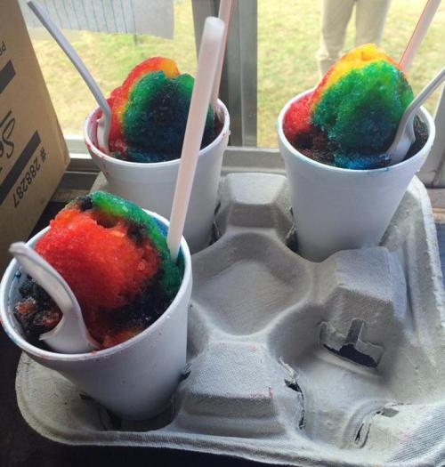 Three colorful snow cones in white cups, sitting in a cardboard holder, with spoons inserted.