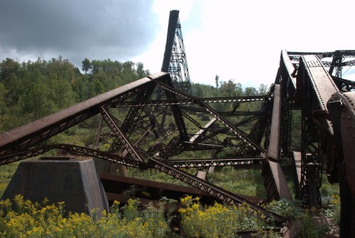 Rusty, abandoned metal structure overgrown with greenery, set against a cloudy sky and distant hills.