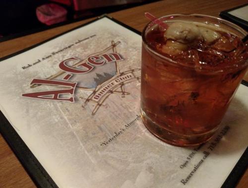 A glass of dark drink with ice sits on a menu at a restaurant, featuring the name "Al-Gen" prominently.