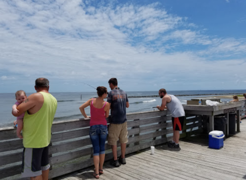 Five people stand on a pier, looking out at the ocean, with a baby in one person's arms and fishing gear nearby.