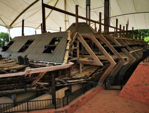 A historic wooden shipwreck displayed under a large canopy, showcasing its weathered structure and cannons.