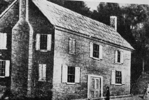 A historic two-story brick house with a chimney, featuring multiple windows and a door, set in a natural landscape.