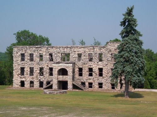 Abandoned stone building with large windows and a staircase, surrounded by grass and a tall tree. Clear blue sky above.