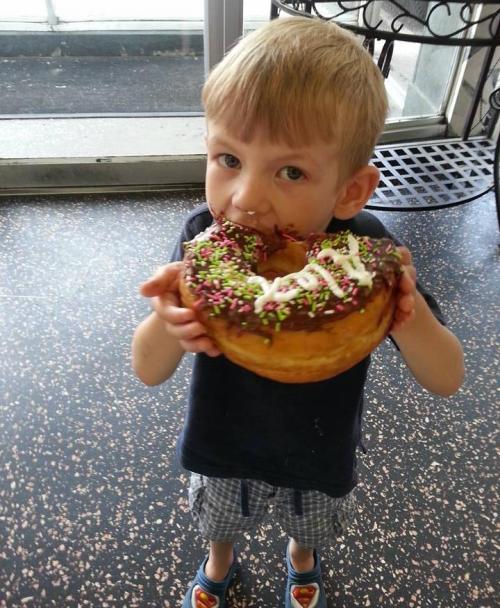 A young boy holds a large chocolate donut topped with sprinkles and icing, smiling happily.