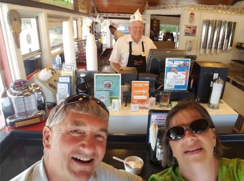 A couple takes a selfie at an ice cream shop, with a smiling server in the background. Bright and cheerful atmosphere.