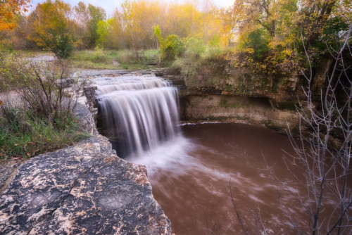 A serene waterfall cascades into a brown pool, surrounded by lush greenery and autumn foliage.