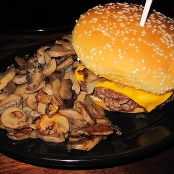 A sesame seed bun burger with melted cheese, accompanied by a side of sautéed mushrooms on a black plate.