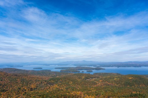 A scenic view of a lake surrounded by colorful autumn foliage and distant mountains under a blue sky.