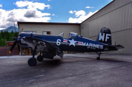 A vintage military aircraft with "MARINES" and "WF" markings parked beside a hangar under a blue sky.