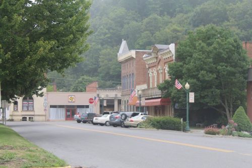 A quiet street scene featuring historic buildings, parked cars, and trees, with American flags displayed.