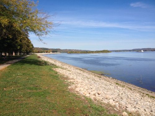 A serene riverside view with a rocky shore, green grass, and trees under a clear blue sky.