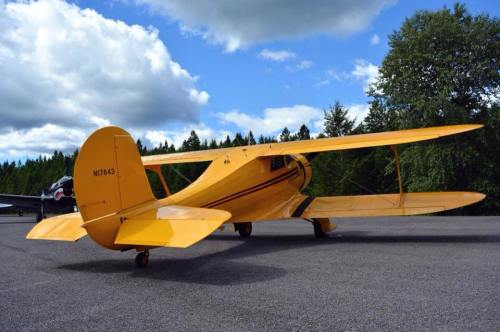 A bright yellow vintage airplane parked on a runway, surrounded by trees and a partly cloudy sky.