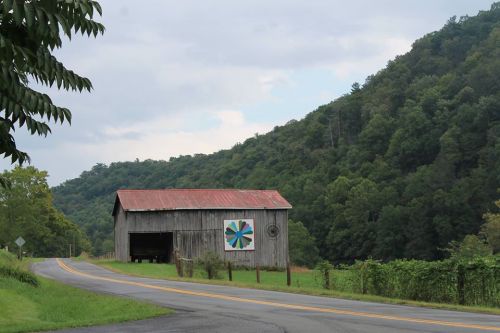 A rustic barn with a colorful wheel design stands beside a winding road, surrounded by lush green hills.