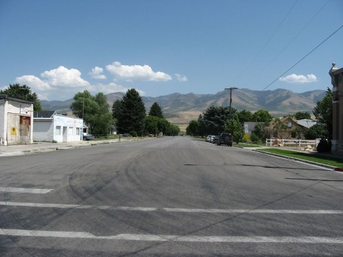A quiet street lined with trees and buildings, with mountains in the background under a clear blue sky.